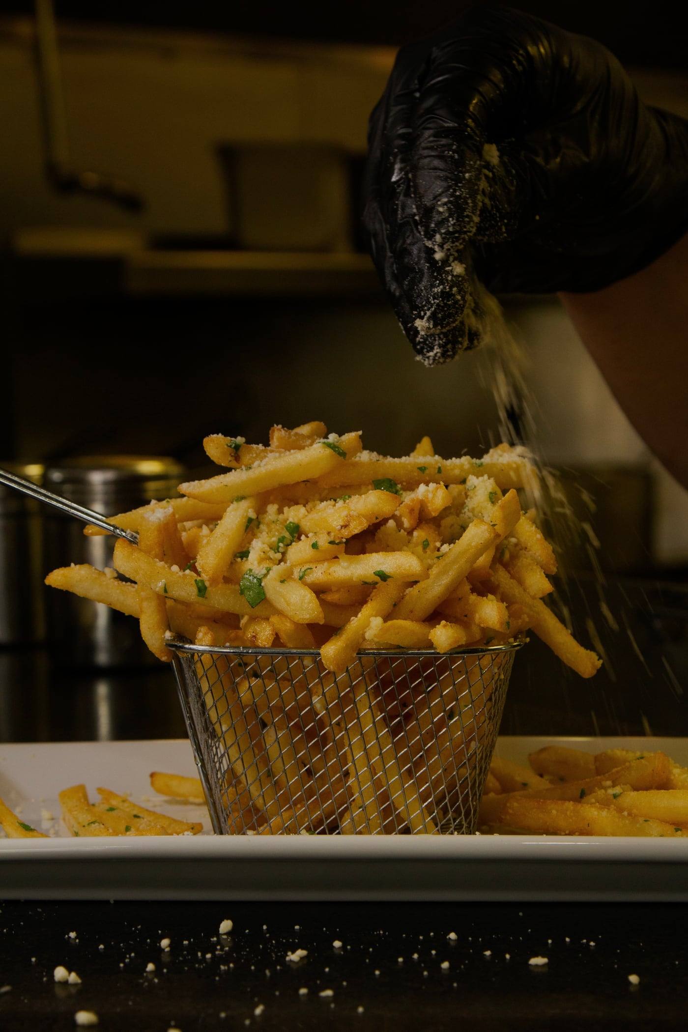A fry basket of golden dirty fries tossed with parmesan and herbs, lifted by a gloved hand in the kitchen